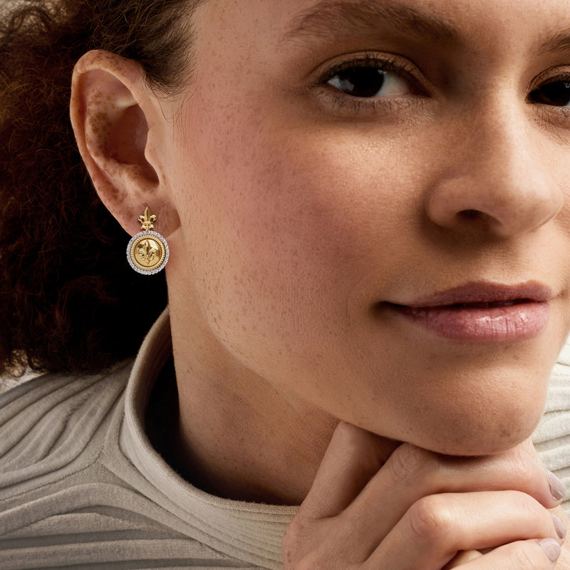 Gold-tone fleur-de-lis drop earring with round diamond-like border, on ear