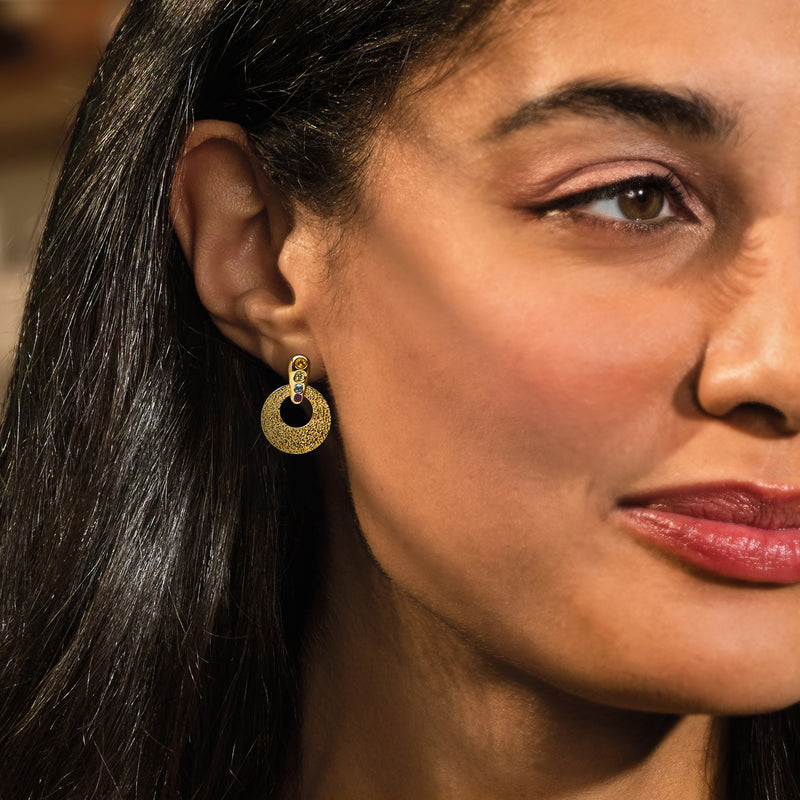 Woman wearing gold-tone open disc drop earrings with bezel-set rainbow gemstones