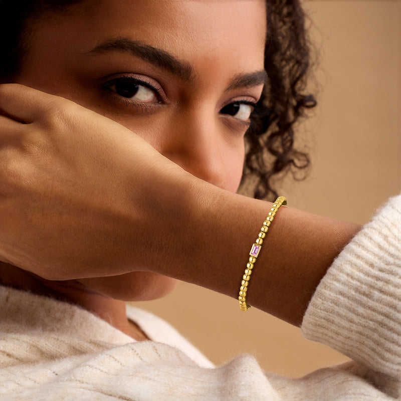 Gold beaded stretch bracelet with pink square sapphire on wrist, close-up.