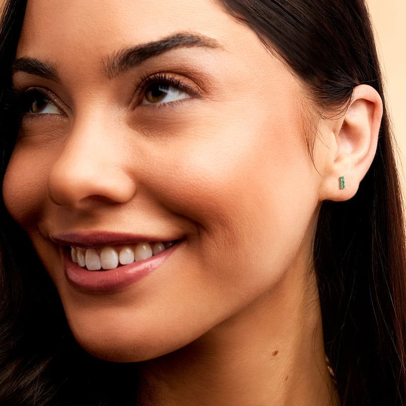 Woman wearing 14kt yellow gold emerald bar stud earrings on ear, green stones visible