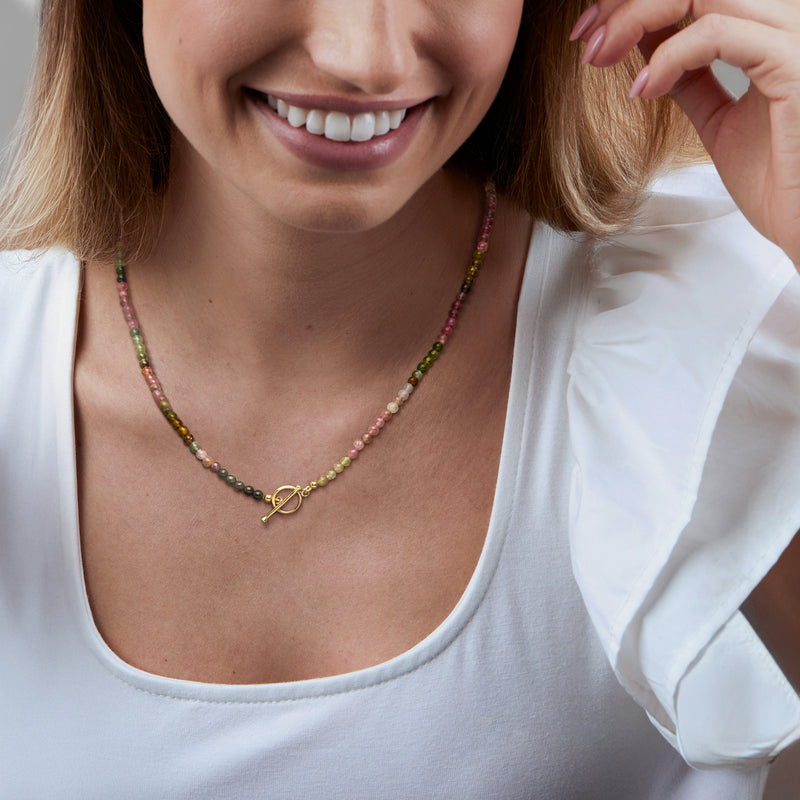 Multicolored tourmaline bead necklace with gold toggle clasp, worn with white top.