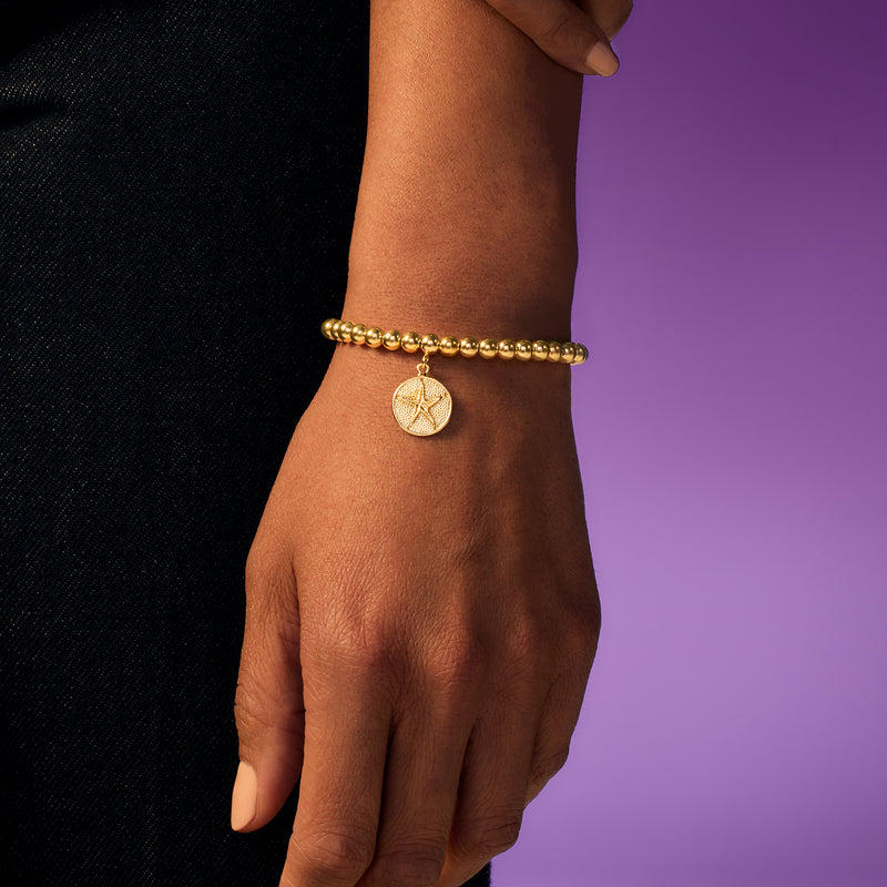 Gold beaded stretch bracelet with sand dollar charm dangling at wrist