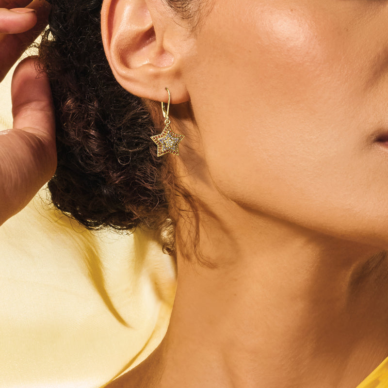 Gold star drop leverback earrings on model’s ear, red and blue stone insets