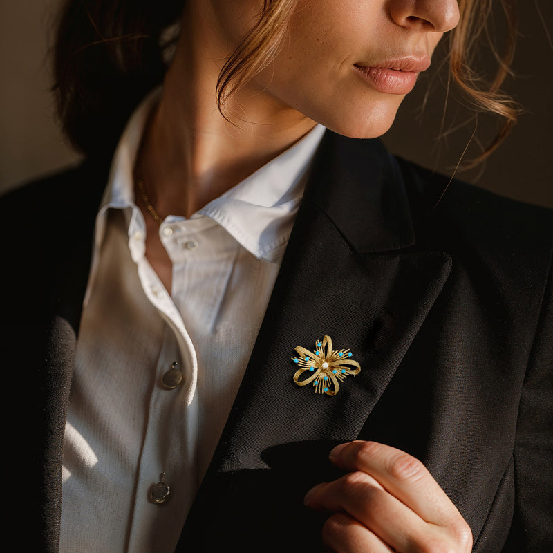 Gold flower ribbon lapel pin with blue stones and white pearl on dark blazer lapel