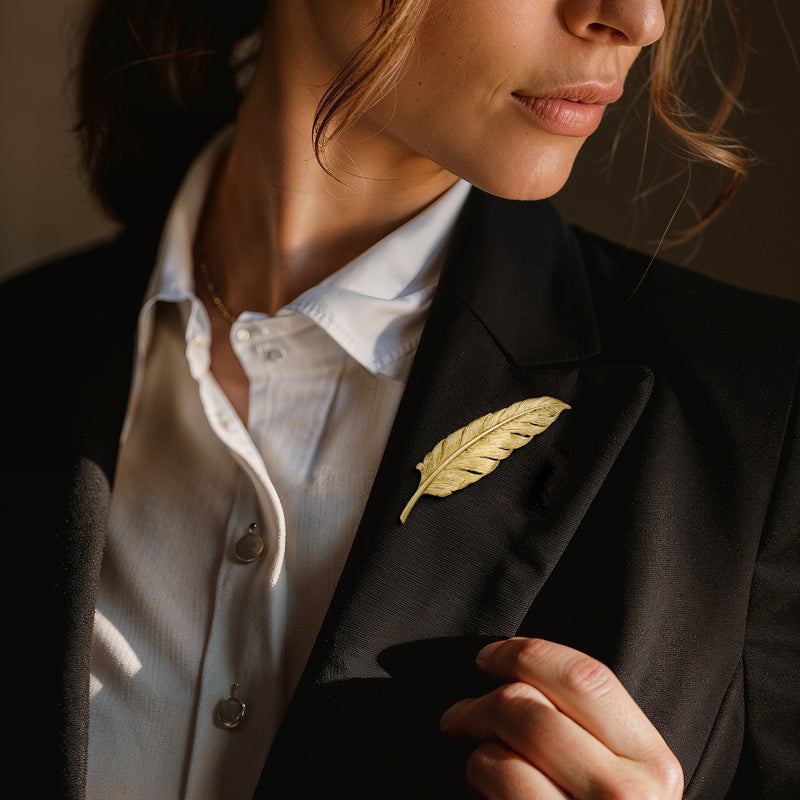 Yellow gold feather lapel pin on a dark blazer jacket near the chest