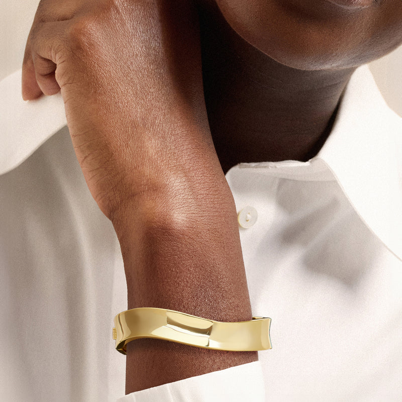 Yellow gold-plated wavy bangle bracelet worn on wrist over shirt sleeve