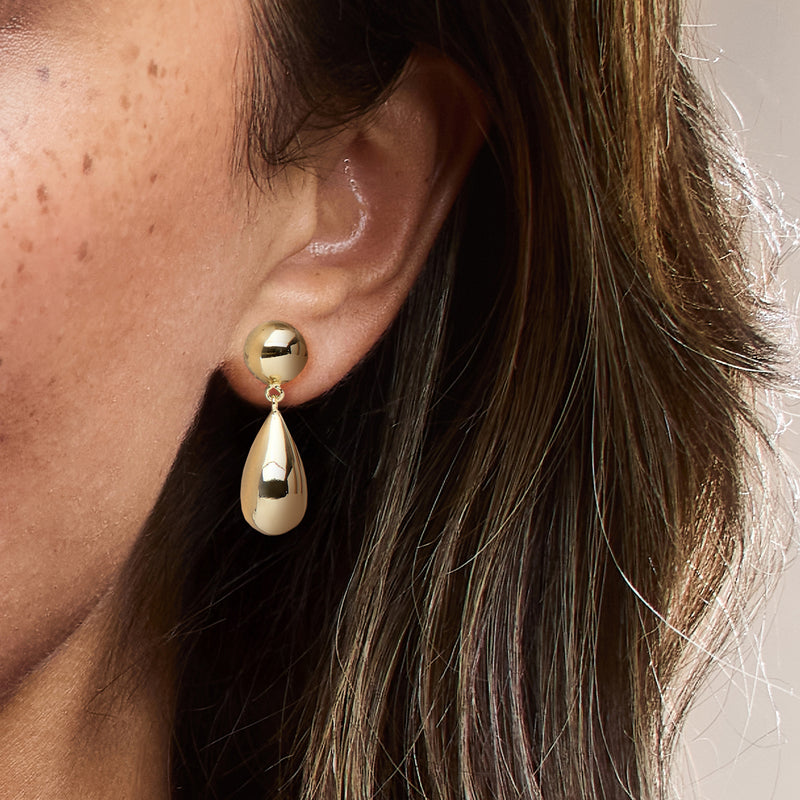Gold-plated teardrop dangle earrings worn, with round top and glossy teardrop drop.
