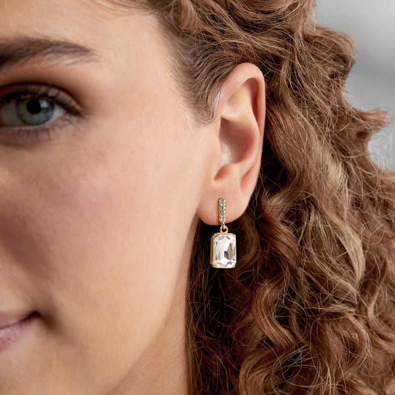 Gold-plated drop earrings with faceted white square gemstone, worn on ear