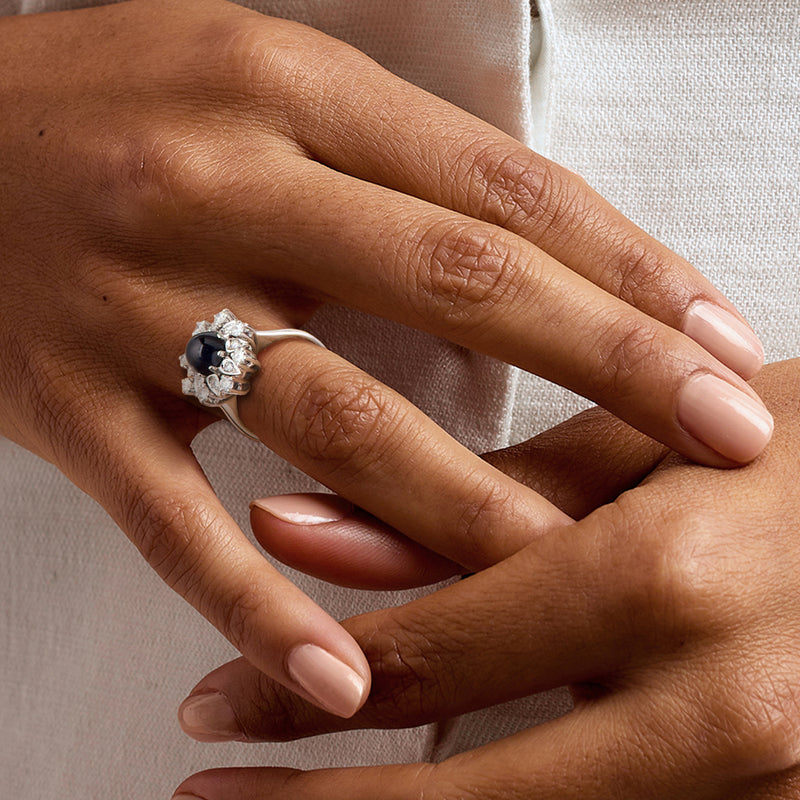 White gold ring with dark blue oval sapphire and round diamond halo, shown on hand