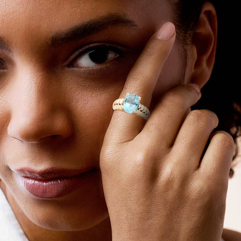 Oval blue topaz ring on finger with ribbed yellow gold band and four-prong setting
