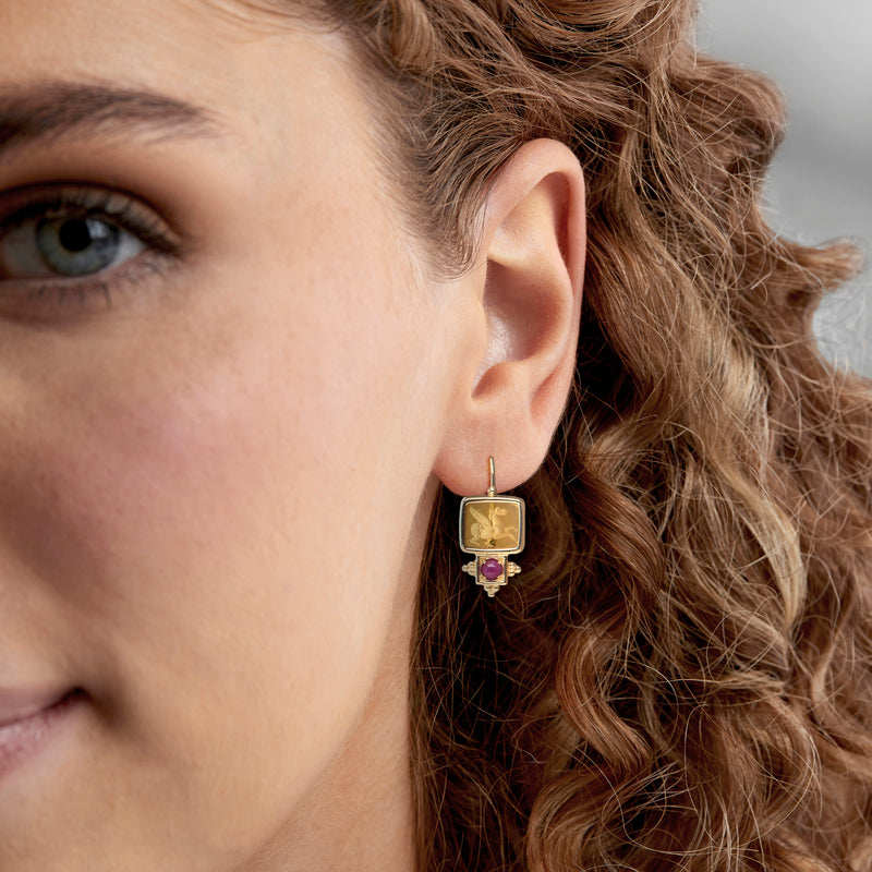 Yellow gold Pegasus earrings with square face and purple ruby, worn on ear