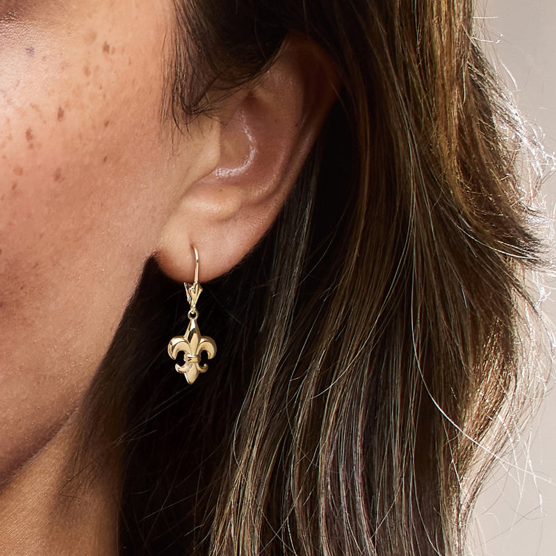 Yellow gold leverback fleur-de-lis drop earrings on ear with small hanging fleur charm