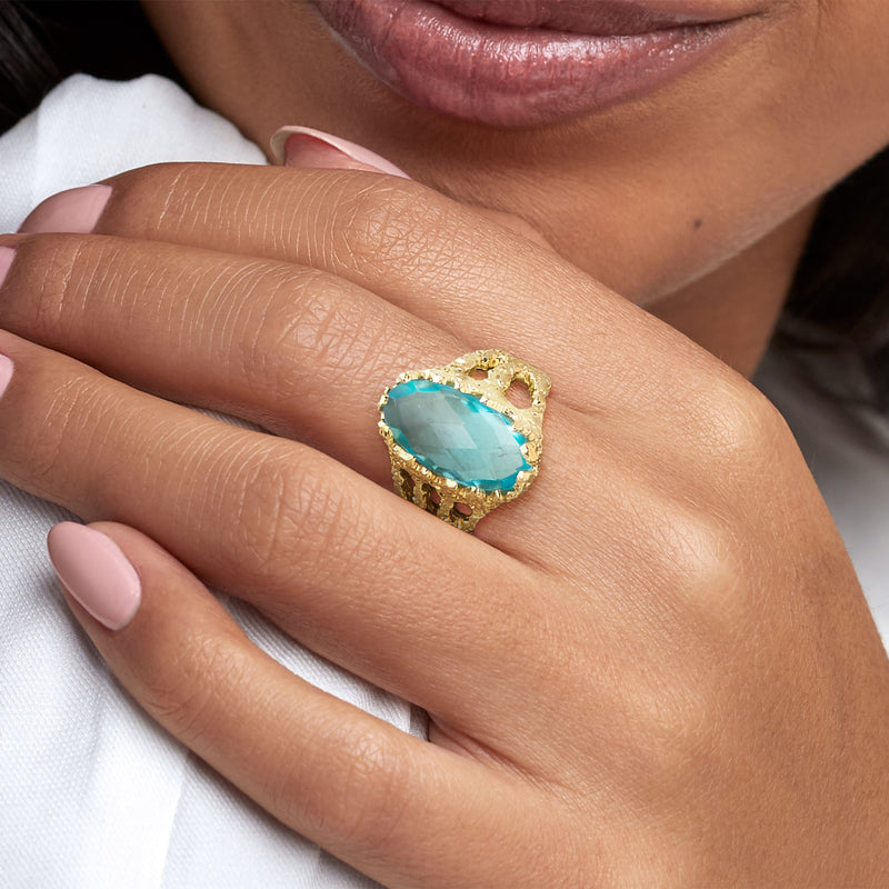 Gold-plated oval blue gemstone ring on finger, showing textured setting