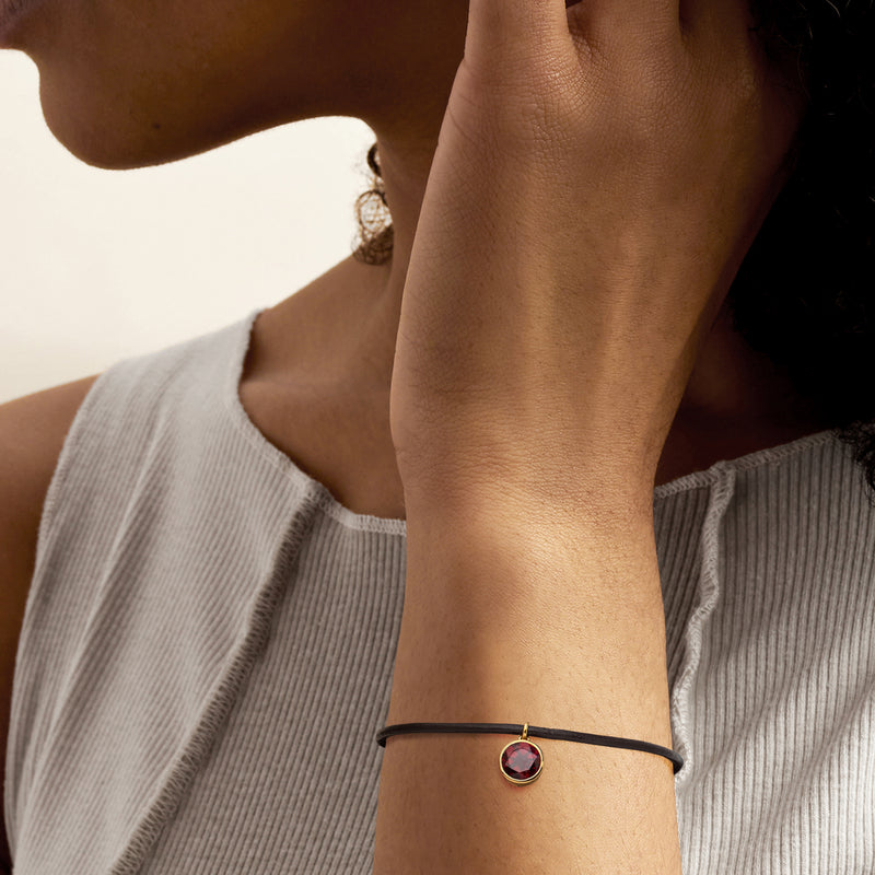 Black leather bracelet on wrist with small round garnet charm and gold-toned clasp