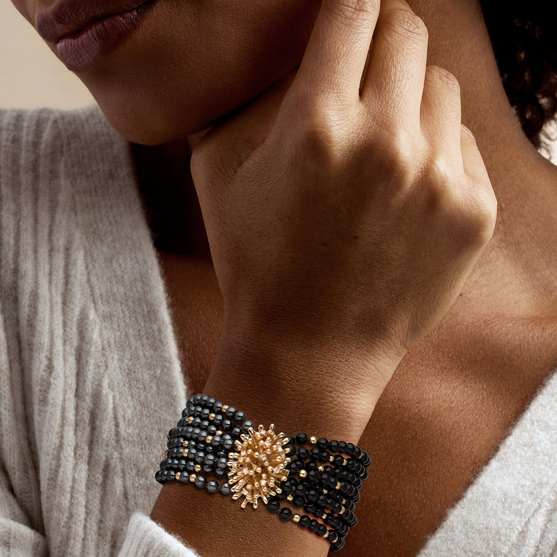 Beaded multi-row bracelet on wrist with dark beads and a gold flowerburst focal charm