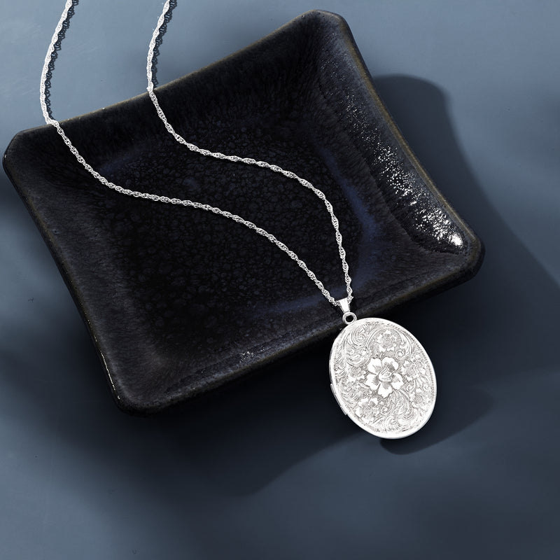 Sterling silver oval locket necklace resting on dark tray, engraved floral front