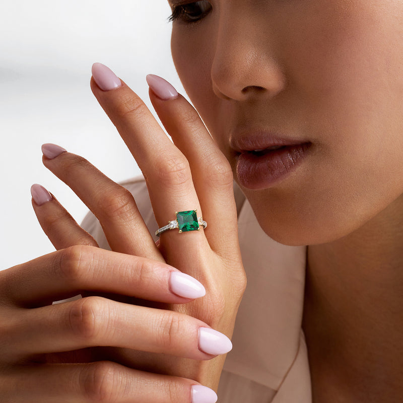 Gold ring with small emerald-cut green stone on model's finger, clear CZ band