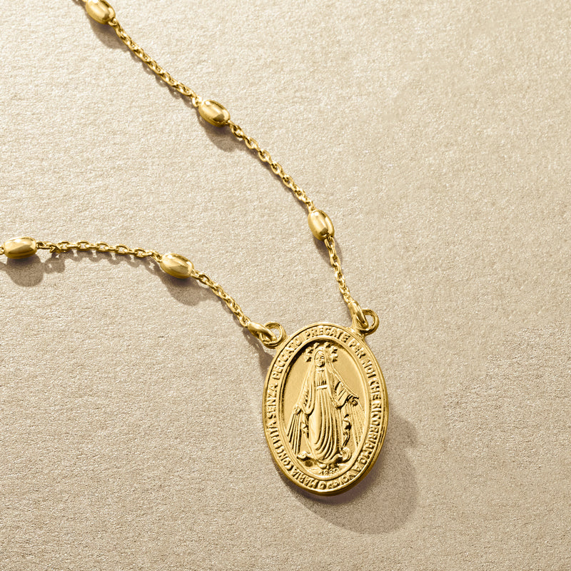 Gold-tone oval Miraculous Medal charm on layered chain resting on light textured surface