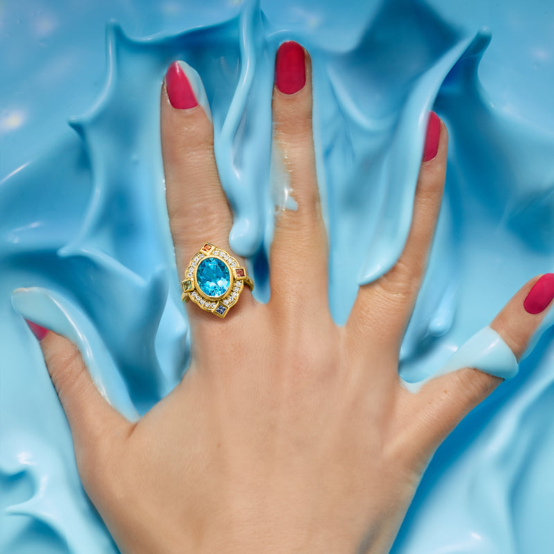 Gold multi-gemstone ring with oval blue center on finger, surrounded by small stones