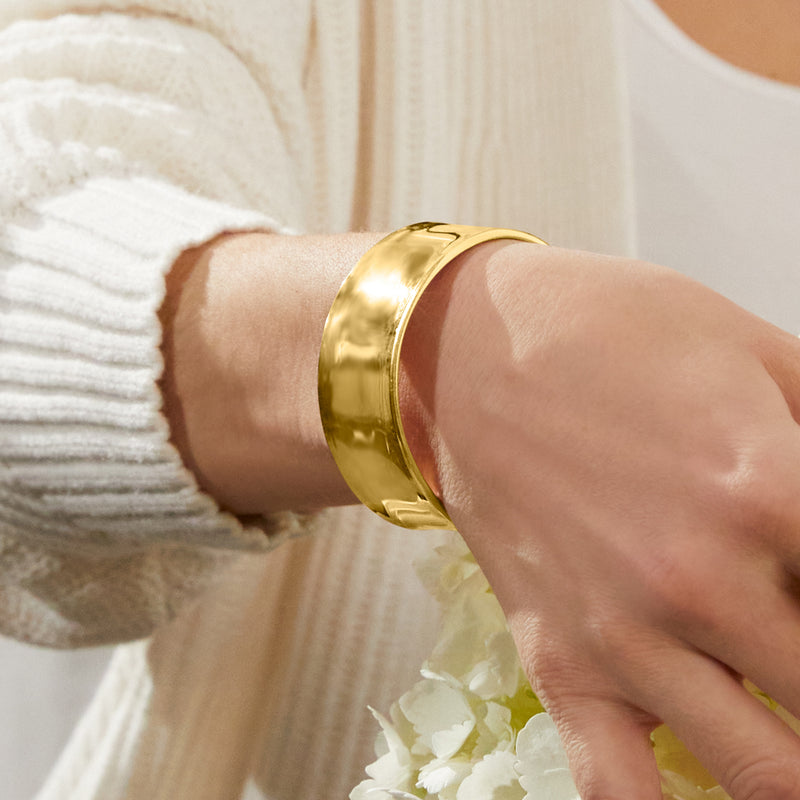 Gold cuff bracelet on wrist over a knit sweater sleeve, smooth polished finish