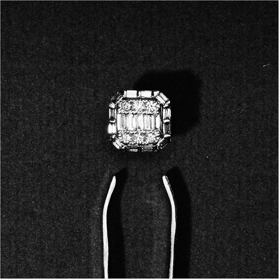 An emerald-cut diamond ring with baguette side stones, held in white metal tweezers against a dark textured background.