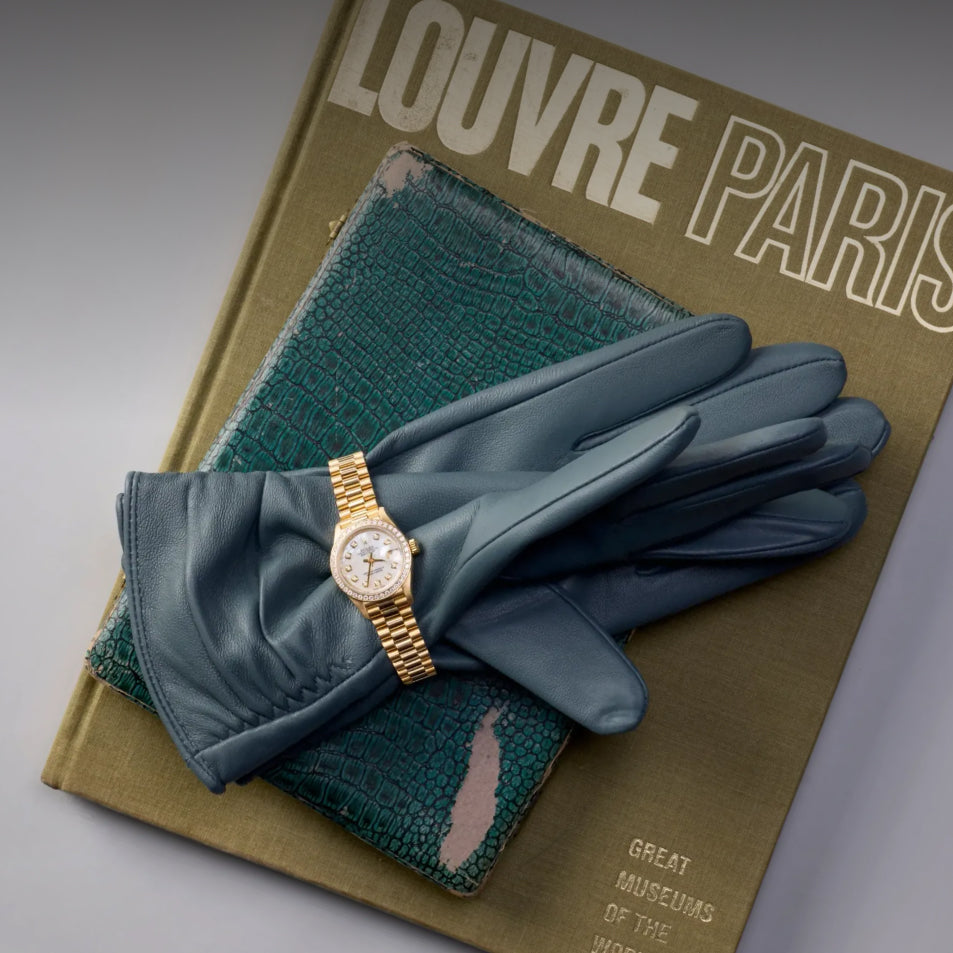 A gold-tone watch with a white dial and bezel is displayed on a pair of slate blue leather gloves, which are arranged on top of a vintage olive-green book titled 'LOUVRE PARIS'.