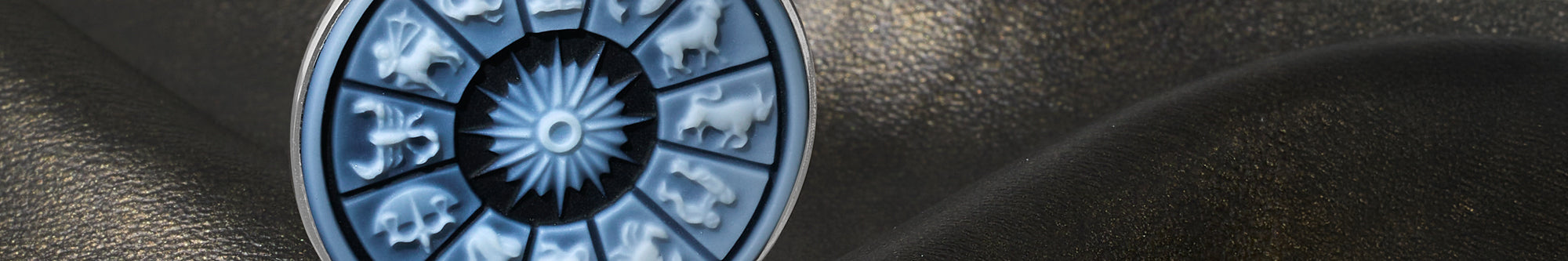 A circular zodiac wheel pendant or medallion with a blue and silver design, featuring the astrological signs around a central sunburst, set against a dark textured background.